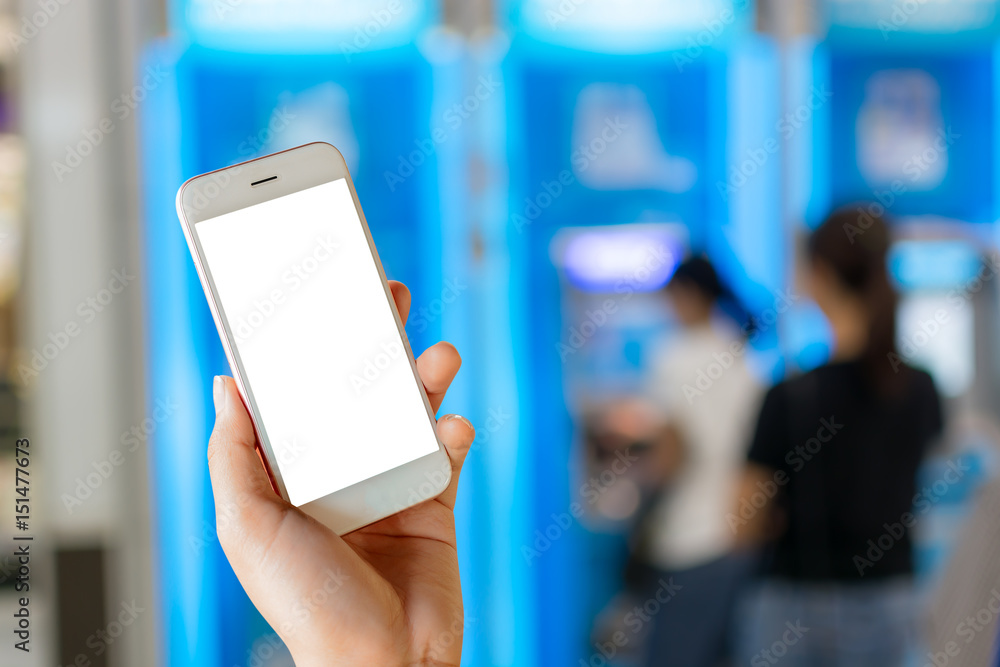 Female hand holding white phone with blank screen on blurred ATM machine background for money financial concept. Female hand holding white phone with blank screen on blurred ATM machine background for money financial concept.