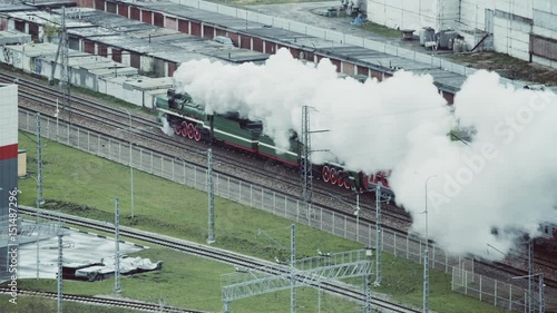 View from distance of several railroad tracks and old retro locomotive blowing a lot of smoke while passing through industrial district, puffer on railway during Victory's day in Moscow, Russia
