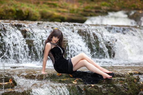 Portrait of a brunette in a black dress near a mountain river. The model is posing at the waterfall.