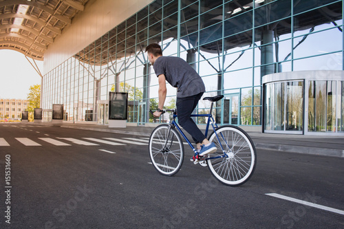  young man riding on bicycle in city street. Man on blue bicycle with white wheels, big mirror windows background