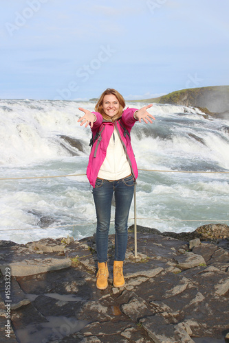 Woman stands in front of the waterfall with outstretched arms and admires beauty of nature