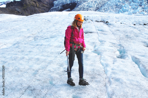 Woman wears pink jacket, crampons and helmet, she stands on the glacier in Iceland and holds axe  