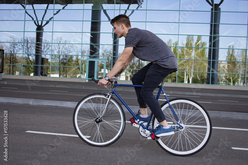 Side view portrait of a young man riding on bicycle in city street. Man on blue bicycle with white wheels, big mirror windows background