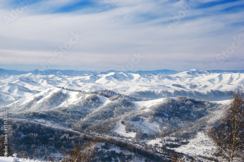 Winter range of snow mountain hills with frost forest under cloudy blue sky. Altai Mountains, Siberia, Russia