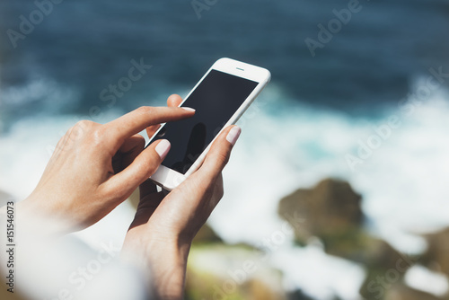 Hipster photograph on smart phone gadget mobile, mock up of blank screen. Traveler hold and using in female hand mobile on background mountain seascape horizon. Tourist look on blue ocean, lifestyle