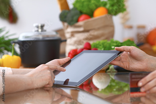 Human hands of two female persons using touchpad in the kitchen. Closeup of two women are making online shopping by tablet computer and credit card. Cooking and shopping concept.