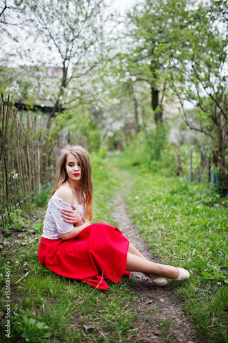 Portrait of sitting beautiful girl with red lips at spring blossom garden on green grass, wear on red dress and white blouse.