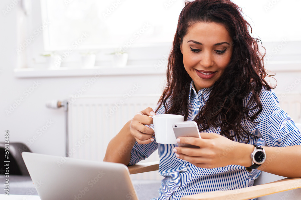 Young woman sitting next to window and texting on phone while drinking morning coffee or tea. Young woman sitting next to window and texting on phone while drinking morning coffee or tea.
