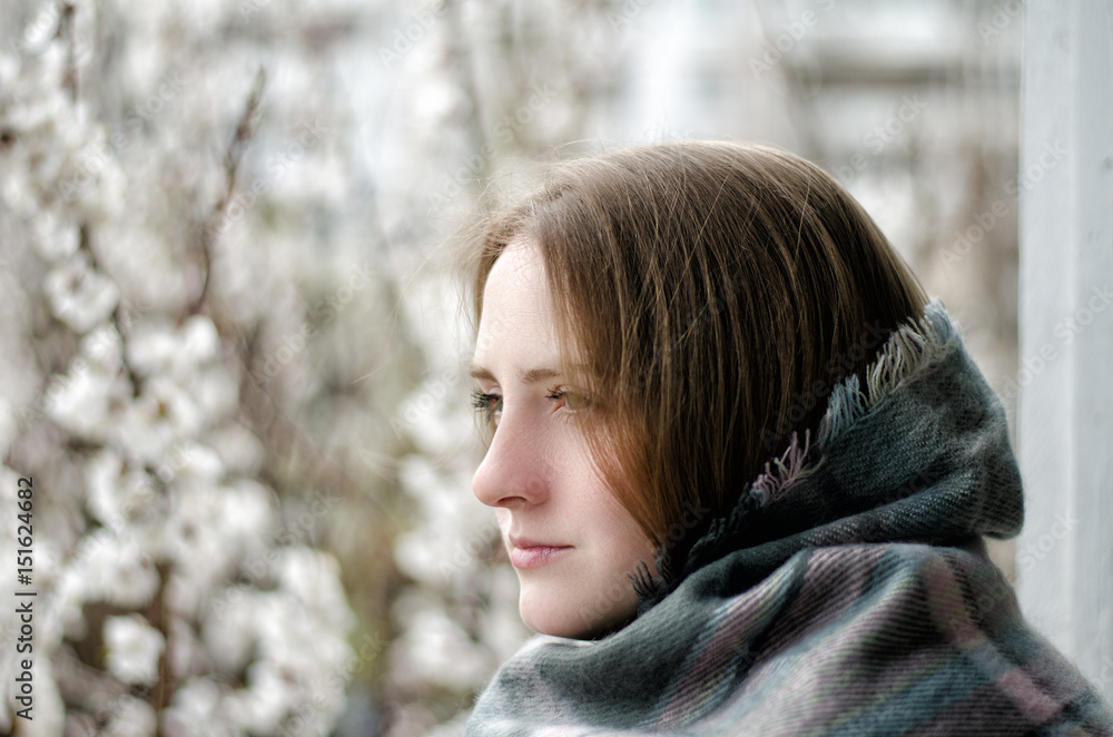 Thoughtful girl, wrapped in a plaid looking into the distance, blossom trees on the background Thoughtful girl, wrapped in a plaid looking into the distance, blossom trees on the background