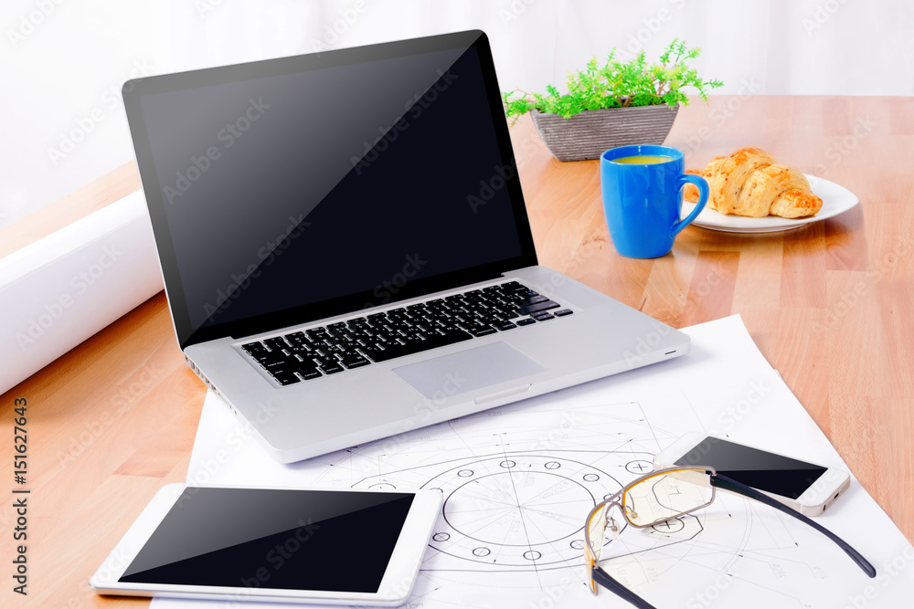 Workplace of cad specialist with laptop, smartphone, tablet, glasses, blue cup and draws on wooden table. Workplace of cad specialist with laptop, smartphone, tablet, glasses, blue cup and draws on wooden table.