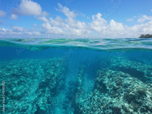 Over under water surface, rocky seabed with coral reef underwater and cloudy blue sky split by waterline, Huahine, Pacific ocean, French Polynesia