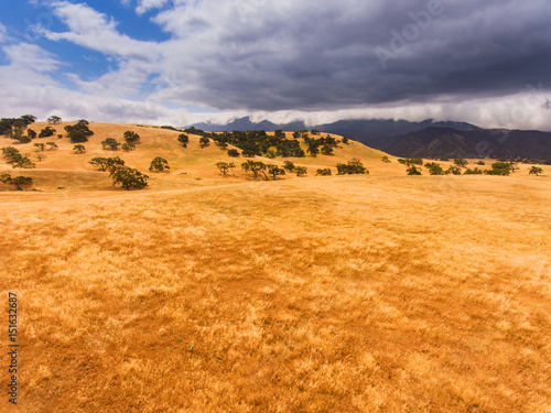aerial of rolling grassy hills with oak trees and stormy mountains