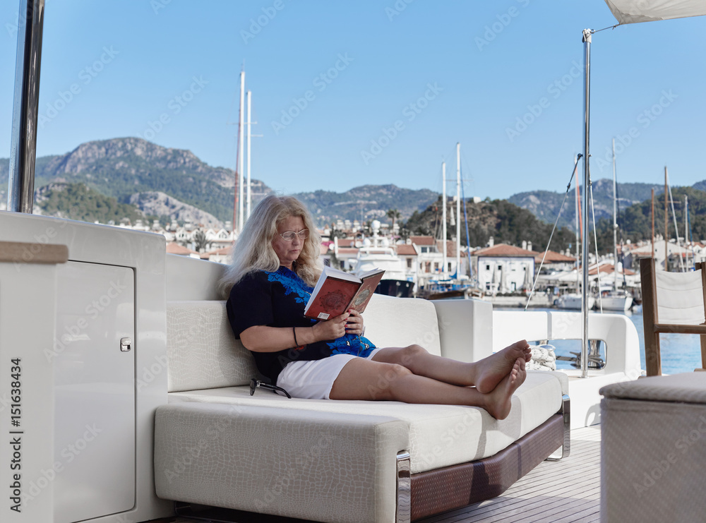 Romantic vacation and travel.  Portrait of good-looking modern blond mature woman wearing glasses, relaxing on the luxury yacht, reading a book. Mediterranean port is on backgroung.