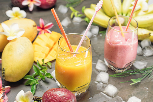 . Mango, banana and strawberry shakes with drinking straw and spoon on black stone table. Passion fruits, frangipani flowers and ice on backgroundTropical fresh smoothie in a glass photo