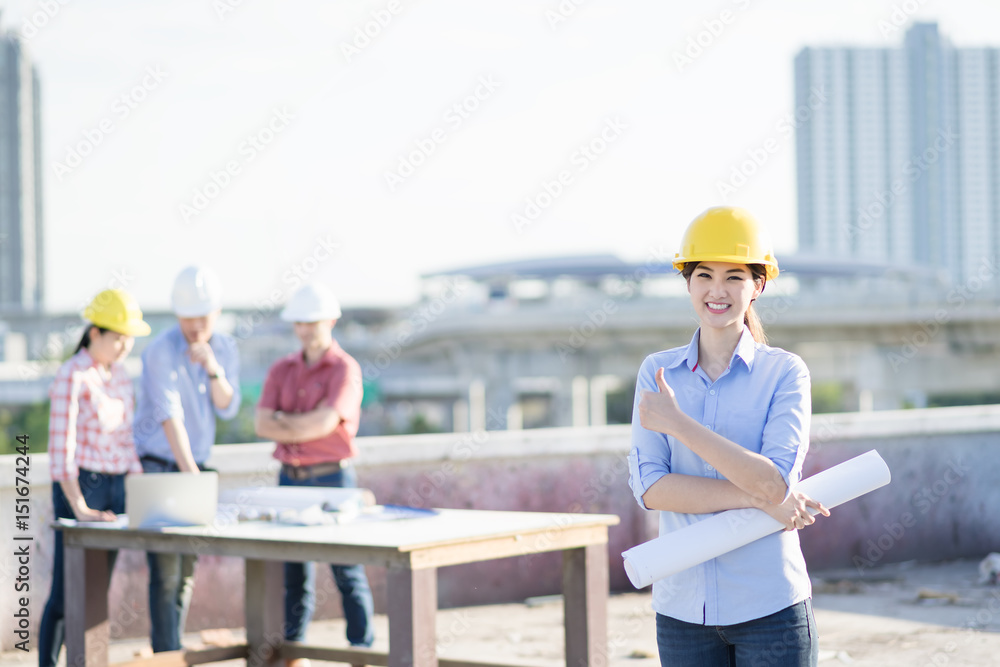 Portrait cute asian woman engineer standing ,thumb up and smiling at construction workplace with team meeting background. Portrait cute asian woman engineer standing ,thumb up and smiling at construction workplace with team meeting background.