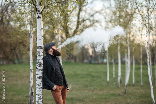 Vape man. Outdoor portrait of a young brutal white guy with large beard and in a vintage cap letting puffs out of steam from an electronic cigarette in the birch grove in the village. Vaping process.