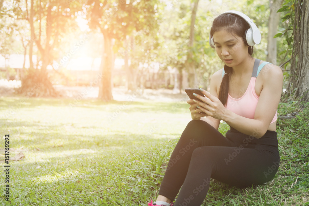 young beautiful asian fitness athlete woman with headphones and smart phone relaxing and listening to music in the city park