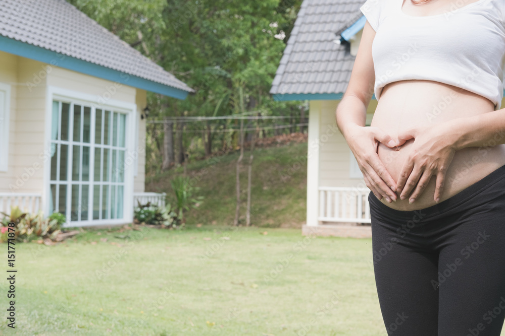 young pregnant woman making heart shape sign on her tummy in front of her house.  pregnancy, maternity belly care concept