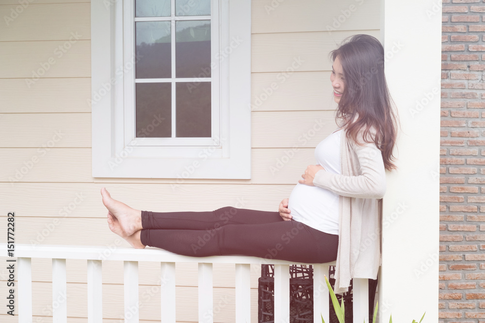 Young asian woman sitting on balcony smiling with pleasure. Beautiful female expecting child having rest in front of her house