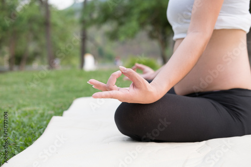 asian pregnant woman practicing yoga while sitting in lotus position on green grass in public park. meditating on maternity. concept of healthy lifestyle and relaxation.