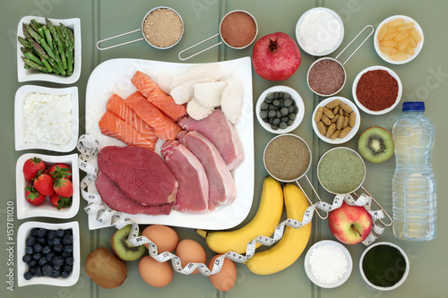 Body building healthy food collection of meat, fish, supplement powders, vitamin pills,fruit, dairy, bottled water and tape measure over slate background.