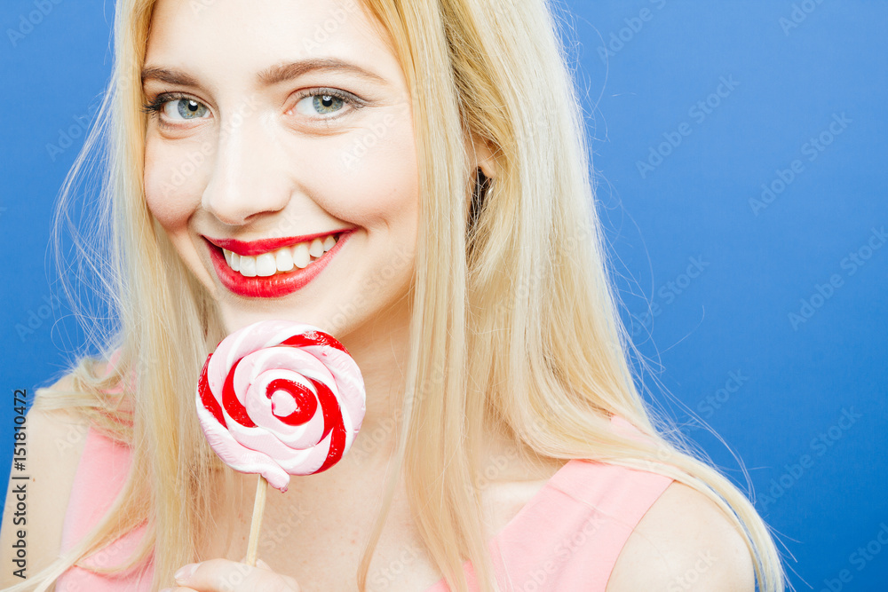 Smiling Pretty Blonde with Sensual Lips and Long Hair is Looking at the Camera Holding Lollipop in Hand on Blue Background in Studio.