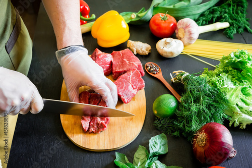 Cook cutting meat on a board and fresh raw vegetables on a dark background. Top view. Flat lay.