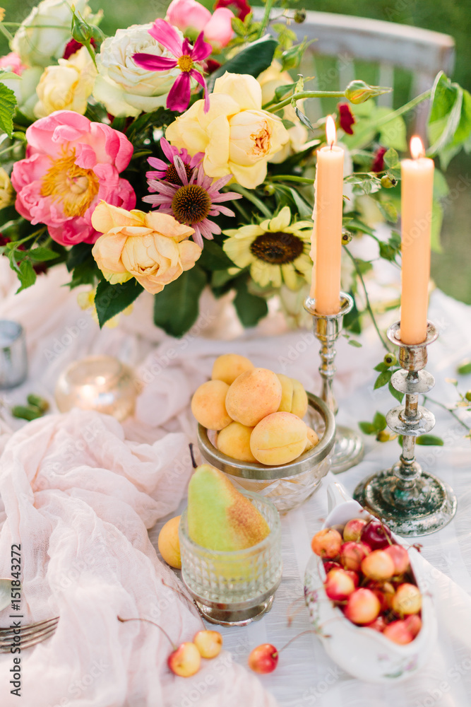 picnic, food, summer, holiday concept - close up on part of the festively decorated table with a beautiful bouquet of roses and peonies, candles in candlesticks, apricots, cherries, pear, glassware