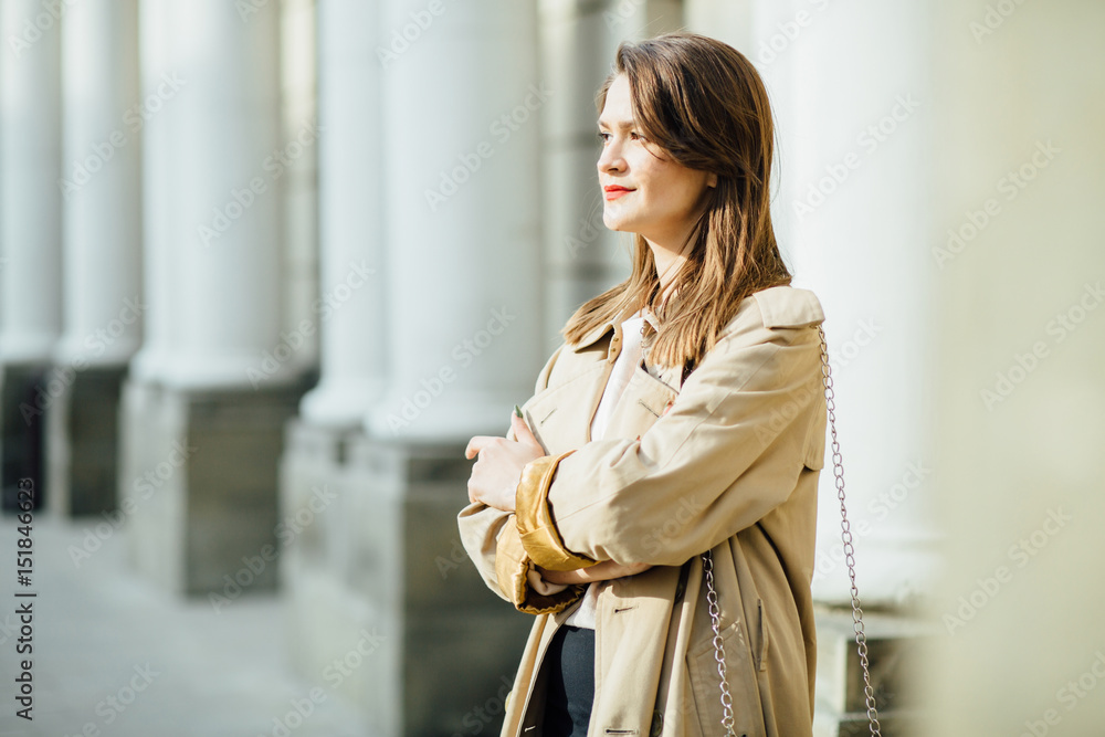 Fototapeta premium Slim elegant woman with red lips in a beige coat and black pants on the white building background