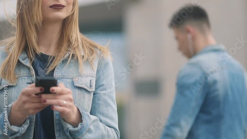 Closeup of amazing youthful girl with blonde hair and dark-red lips girl inblue denim court and strict bluse, communicating with someone via smartphone on the urban city street photo