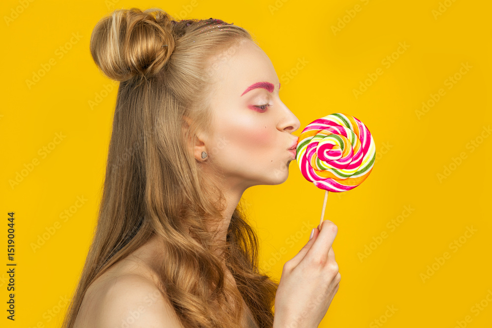 Bright portrait of young funny beautiful woman with colorful lollipop over yellow background.