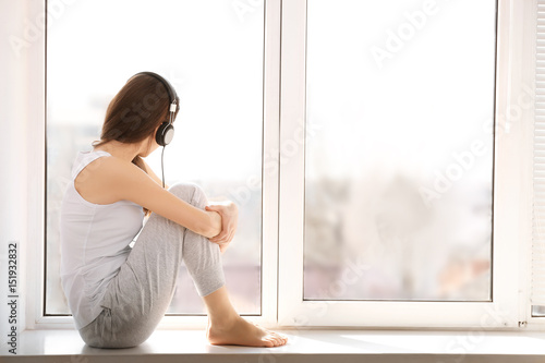 Beautiful young woman sitting on windowsill and listening to music