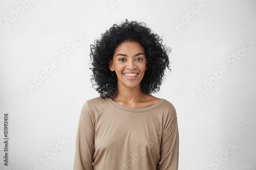 Waist up portrait of cheerful young mixed race female with curly hair posing in studio with happy smile. Dark-skinned woman dressed casually smiling joyfully, showing her white straight teeth photo