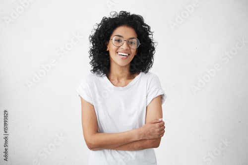 Charming young dark-skinned woman with curly hairstyle having shy smile posing in studio in closed posture, keeping arms folded, feeling constrained and a bit nervous. Human emotions and feelings photo