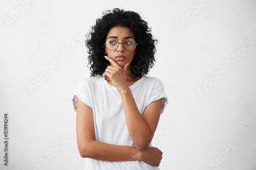Portrait of beautiful casually dressed young woman in round glasses having doubtful expression, looking away in indecisiveness, holding her chin, trying to find best solution. Body language © wayhome.studio 
