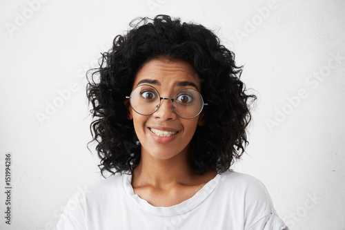 Headshot of funny young mixed race female with curly hair wearing stylish eyeglasses looking at camera with beseeching or shocked expression, raising her eyebrows. Human emotions and feelings