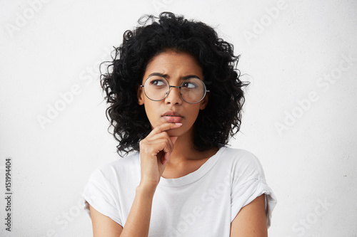 Isolated portrait of stylish young mixed race woman with dark shaggy hair touching her chin and looking sideways with doubtful and sceptical expression, suspecting her boyfriend of lying to her photo