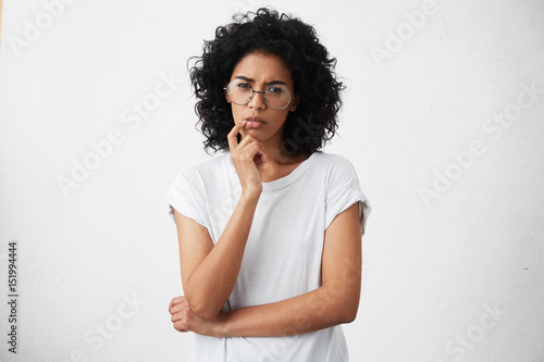 Human emotions, feelings, reaction and attitude. Attractive girl in casual t-shirt and round glasses squeezing eyes and keeping hand on chin in doubt and suspicion, feeling sceptical about something photo
