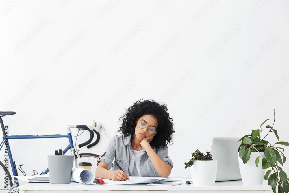 Stylish mixed race student designer with curly hairstyle fell asleep while doing home work in her white room, feeling exhausted as she has to finish project as soon as possible because of deadline