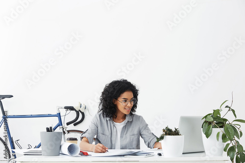Happy young dark-skinned woman architect wearing round eyeglasses and grey shirt smiling, making drawing with pencil and checking measured data on laptop computer screen, enjoying her profession
