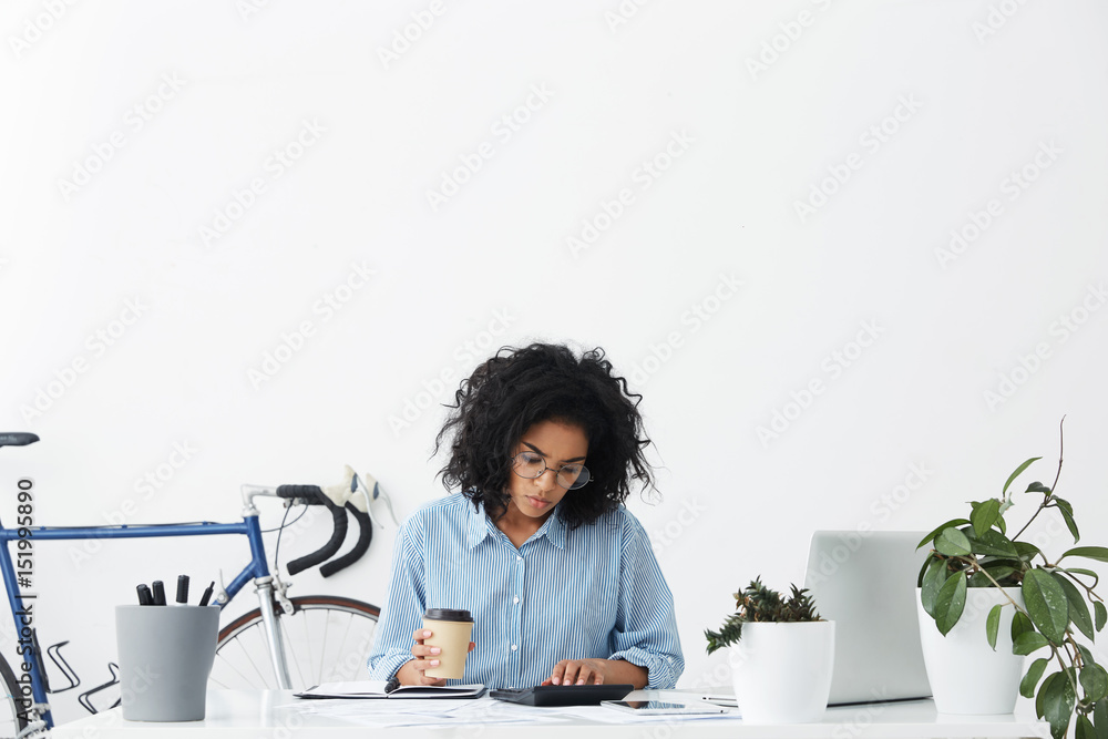 Concentrated hardworking graduated student girl wearing blue shirt and glasses having another coffee out of paper cup while working on diploma project at her workplace, checking calculations Concentrated hardworking graduated student girl wearing blue shirt and glasses having another coffee out of paper cup while working on diploma project at her workplace, checking calculations