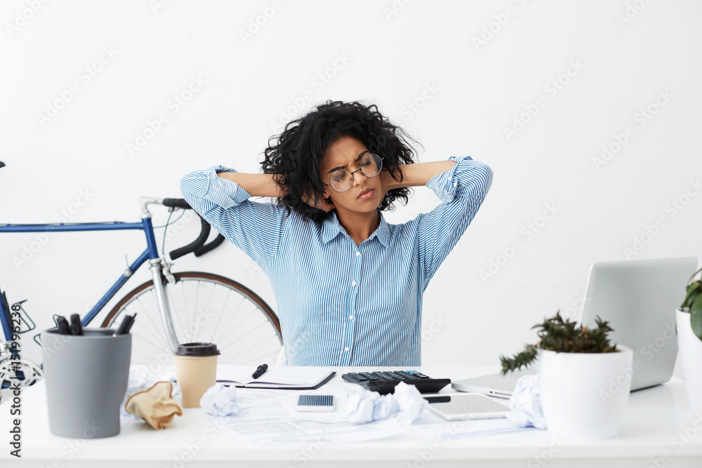 Portrait of black student stretching arms being tired after finishing her homework, feeling exhausted after difficult math task, sitting in front of table with calculator, tablet, coffee and notebook Portrait of black student stretching arms being tired after finishing her homework, feeling exhausted after difficult math task, sitting in front of table with calculator, tablet, coffee and notebook