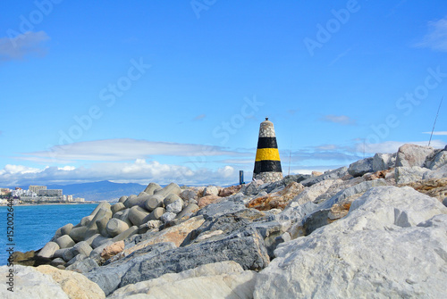 A view to Mediterranean sea, a lighthouse with breakwaters, fishing rods of locals and Torremolinos at the background from a pier at Benalmadena port Puerto Marina, Andalusia, Spain. photo