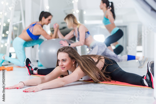 Fit young women working out in gym with smiling young girl in focus doing full split leaning forward