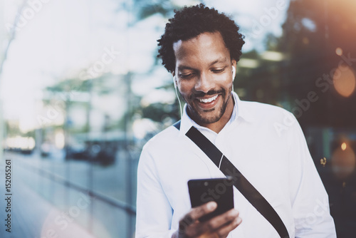 Happy young African American man in headphone walking at sunny city and making video conversation with friends on his electronic smart phone.Blurred background.
