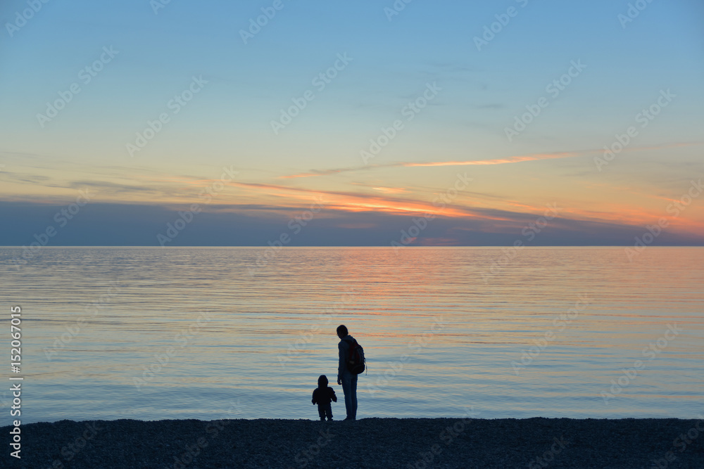 woman with backpack and small child standing on beach and watching sunset at sea woman with backpack and small child standing on beach and watching sunset at sea