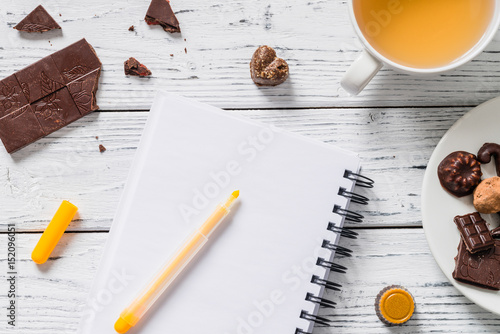 Blank notebook with cup of tea and raw gluten free chocolate candies. Business or lifestyle morning concept on white wooden background. Top view with copy space