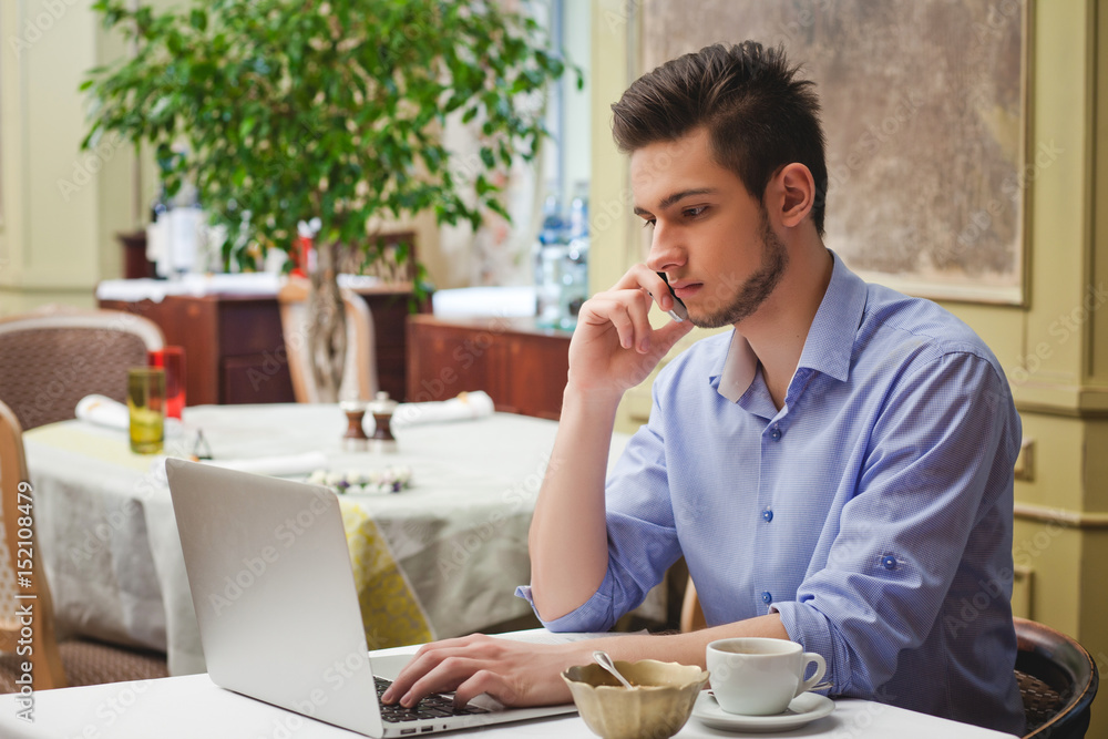 Young guy sitting in cafe with laptop, talking by phone and drin Young guy sitting in cafe with laptop, talking by phone and drin