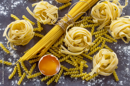 Different kinds of pasta spaghetti, fusilli, fettuccine and raw egg yolk on the kitchen wooden table.