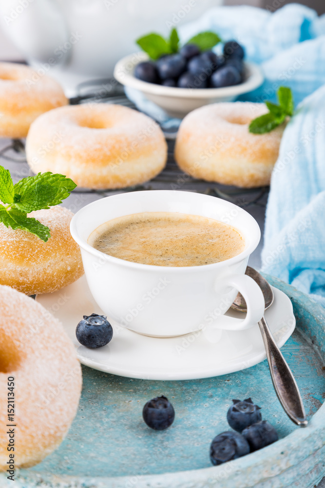 Cup of coffee and homemade donuts with powdered sugar and fresh blueberries on light gray background. Selective focus. Cup of coffee and homemade donuts with powdered sugar and fresh blueberries on light gray background. Selective focus.